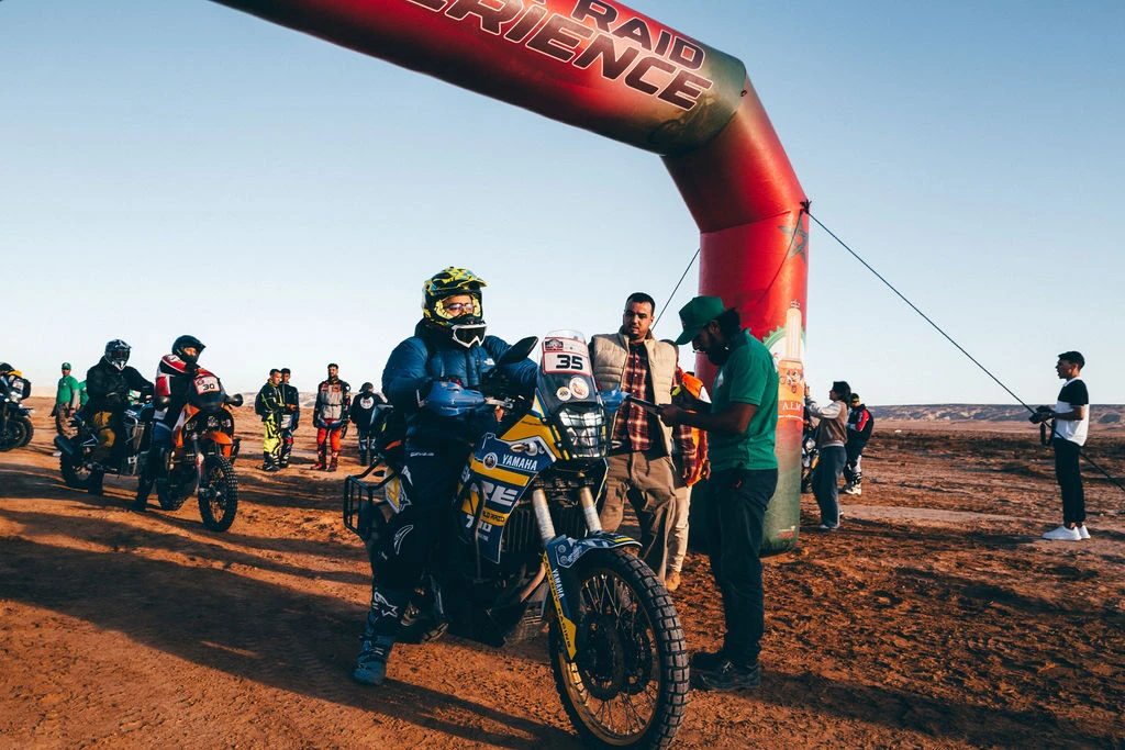 Riders and bikes gathered at the Sahara Raid Experience start arch on desert ground in Morocco