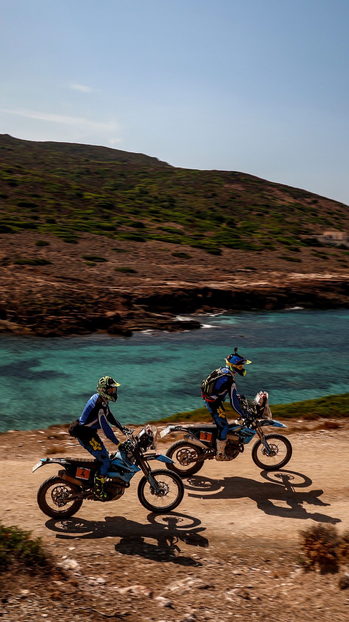Two riders climbing a coastal gravel track above turquoise water during Swank Rally di Sardegna